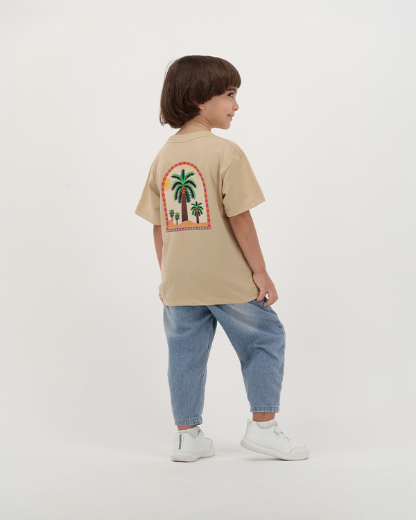 Boy wearing beige The Palm Kids Leaf Print T-Shirt with palm tree design on back.